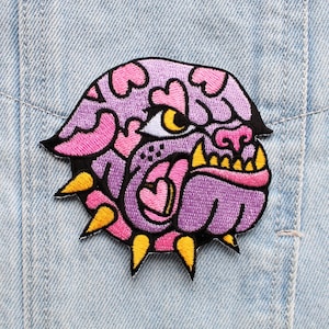 Może przedstawiać: Haftowany naszywka w kolorze fioletowym i różowym przedstawiająca buldoga z kolczastym kołnierzem i sercami. Buldog ma żółte oko i różowy język.