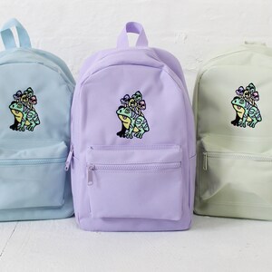 Könnte beinhalten: Drei pastellfarbene Rucksäcke mit einem gestickten Frosch- und Pilzmotiv auf der Vorderseite. Die Rucksäcke sind hellblau, lavendelfarben und hellgrün.