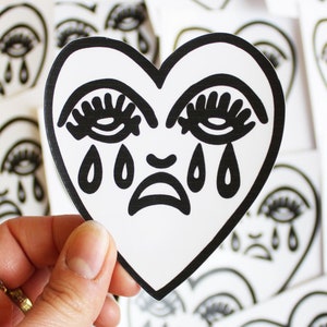 May include: A black and white sticker of a crying heart. The heart has a sad face with eyelashes and tears. The sticker is on a white background.