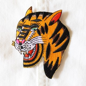 May include: Embroidered tiger patch with orange, black, pink, and green details. The tiger has an open mouth, showing red and white teeth. The patch is on a white background.