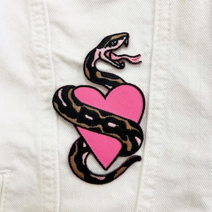 May include: Embroidered patch featuring a pink heart entwined by a black and brown snake. The snake has a detailed head with a forked pink tongue. The patch is likely designed to be ironed or sewn onto fabric, adding a decorative element to clothing or accessories.