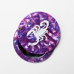 May include: A round, purple and pink compact mirror with a silver scorpion design. The compact is open, revealing a mirror. The compact has a textured, marbled appearance and a small gold-colored hinge.