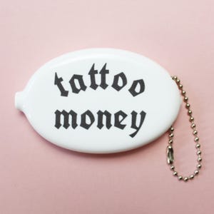 May include: White oval coin purse with the words "tattoo money" in black gothic font. The purse has a small silver chain attached to it. The background is a solid pink color.
