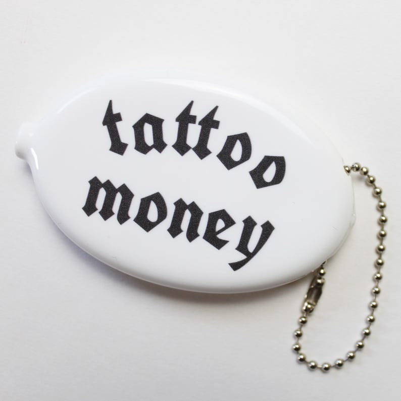 May include: White oval coin purse with the words "tattoo money" printed in black gothic font. The purse has a small metal ball chain attached to it.