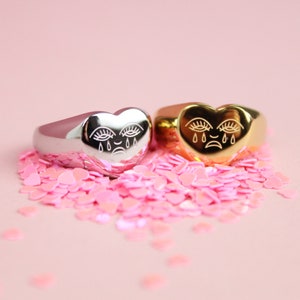 May include: Two heart-shaped rings with a crying face design. One ring is silver and the other is gold. The rings are surrounded by pink heart-shaped confetti.