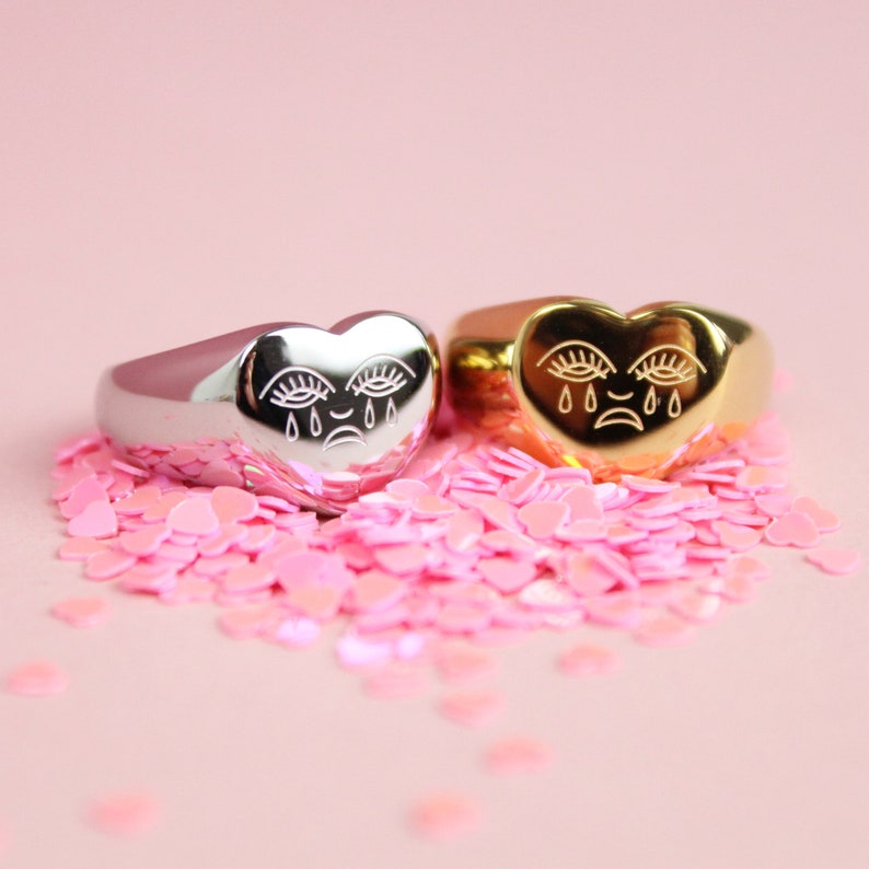 May include: Two heart-shaped rings with a crying face design. One ring is silver and the other is gold. The rings are surrounded by pink heart-shaped confetti.