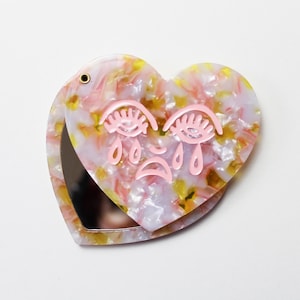 Pode incluir: Espelho compacto em forma de coração com um design marmorizado rosa e amarelo. O espelho apresenta um design de rosto chorando rosa na frente. O compacto está aberto, revelando uma superfície reflexiva.