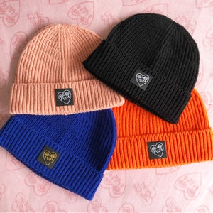 Könnte beinhalten: Vier Strickmützen in verschiedenen Farben: rosa, schwarz, blau und orange. Jede Mütze hat einen kleinen gestickten Aufnäher mit einem Herzdesign und einem weinenden Gesicht.