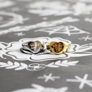 May include: Two heart-shaped rings with a crying face design. One ring is silver and the other is gold.