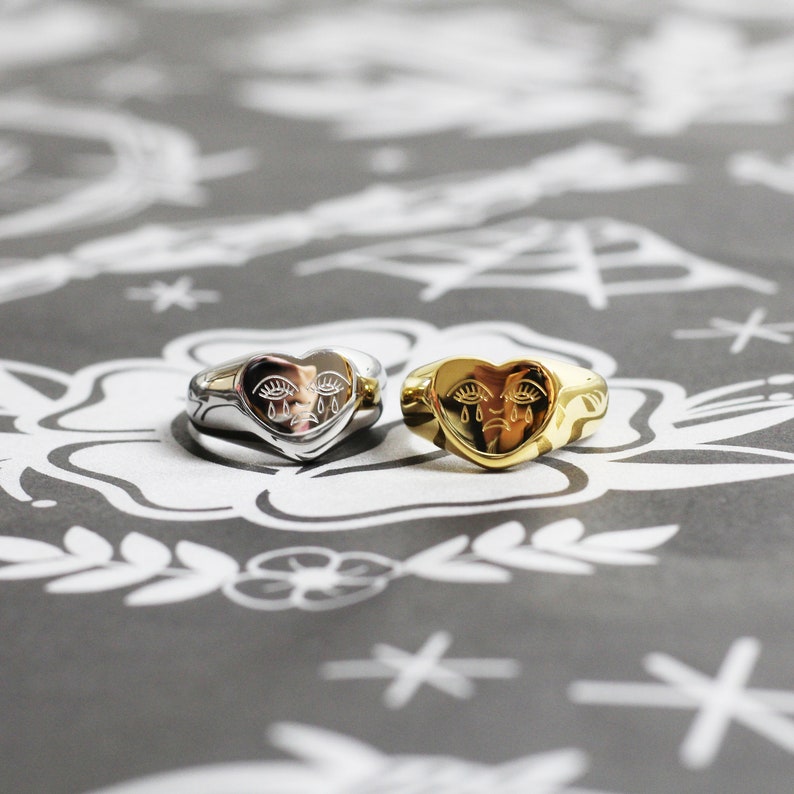 May include: Two heart-shaped rings with a crying face design. One ring is silver and the other is gold.