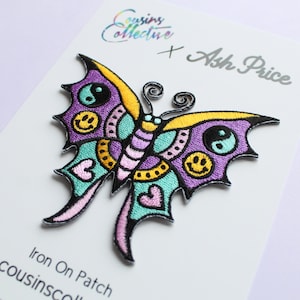 May include: Iron-on patch featuring a purple butterfly with yellow, green, and pink accents. The butterfly has a yin-yang symbol, smiley faces, and hearts on its wings. The patch is labeled "Iron On Patch" and "cousinsco".