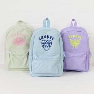 May include: Three pastel colored backpacks with embroidered designs. The light green backpack has a pink rose and the text "Aubrey". The light blue backpack has a blue heart with a crying face and the text "Casper". The purple backpack has a yellow butterfly and the text "Daphne".