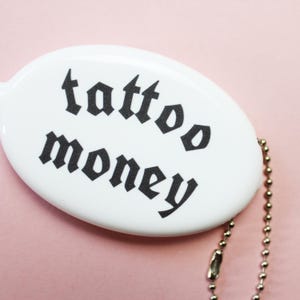 May include: White oval keychain with the words "tattoo money" in black gothic font. The keychain has a silver ball chain attached. The background is a solid light pink.