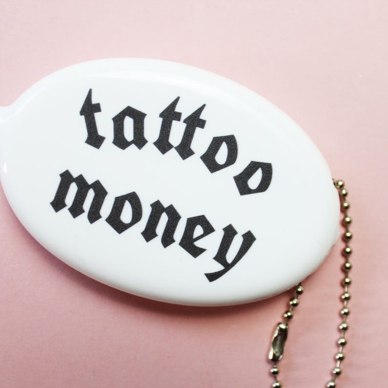 May include: White oval keychain with the words "tattoo money" in black gothic font. The keychain has a silver ball chain attached. The background is a solid light pink.