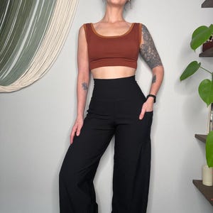 May include: A woman wearing a rust-colored crop top with a brown trim and high-waisted black wide-leg pants. The pants have pockets. The outfit is simple and stylish.