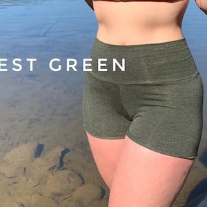 May include: Forest green high-waisted shorts made from a soft, stretchy fabric. The shorts are shown on a person standing in front of a body of water.