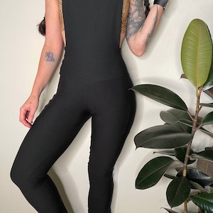 May include: Black jumpsuit with wide straps and flared legs. The back of the jumpsuit has a patterned design in shades of orange and brown. The jumpsuit is made of a stretchy material and has a fitted silhouette.