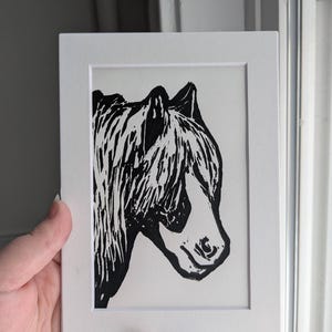 May include: Black and white linocut print of a horse's head, framed in a white mat. The artwork features a detailed depiction of the horse's profile, with a focus on the mane and facial features. The print is held against a neutral background.