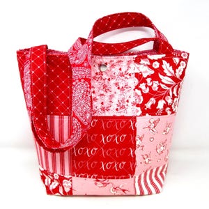 May include: A patchwork tote bag in shades of red and pink. The bag features various fabric patterns, including floral, stripes, and a paisley design. The front of the bag has the text "xoxo" in a script font. The bag has two handles.