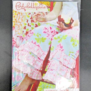 May include: A sewing pattern packet titled Portabellopixie Boutique Patterns for ruffle pants by Sandi Henderson. The cover features a photograph of light blue pants with pink floral patterns and three tiers of white ruffled hems.