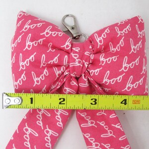 May include: A pink fabric bow with the word "boo" in white script repeated across the surface. The bow measures approximately 5 inches across and has a silver-colored metal clasp.