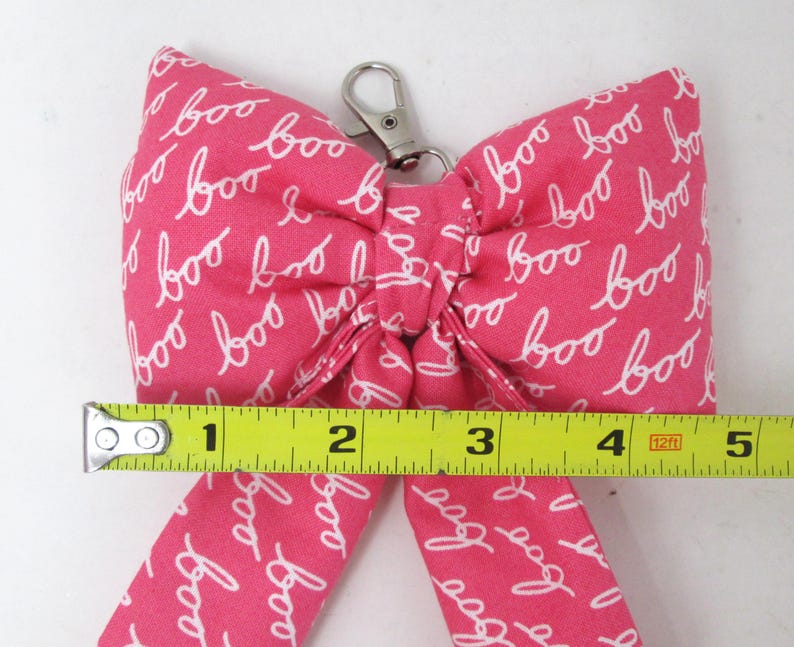 May include: A pink fabric bow with the word "boo" in white script repeated across the surface. The bow measures approximately 5 inches across and has a silver-colored metal clasp.