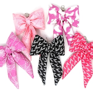 May include: Five fabric bows in various Halloween-themed patterns. The bows feature designs like spiderwebs, ghosts, and the word "boo." Colors include pink, black, and white. Each bow has a metal clip.