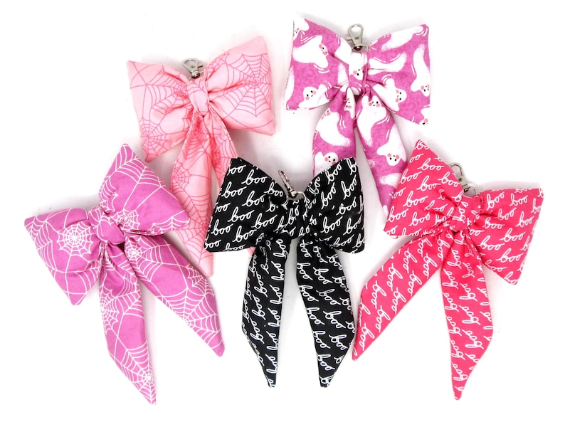 May include: Five fabric bows in various Halloween-themed patterns. The bows feature designs like spiderwebs, ghosts, and the word "boo." Colors include pink, black, and white. Each bow has a metal clip.