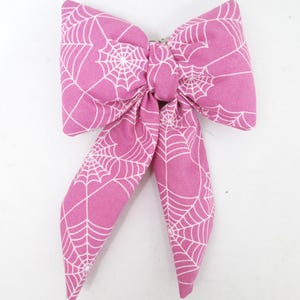 May include: A pink fabric bow with a white spiderweb pattern. The bow is tied in the center and has two long, flowing tails. The bow is likely used as a hair accessory or decoration.