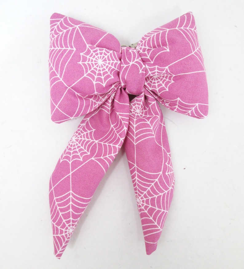 May include: A pink fabric bow with a white spiderweb pattern. The bow is tied in the center and has two long, flowing tails. The bow is likely used as a hair accessory or decoration.