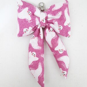 May include: A pink bow-shaped accessory with a white ghost pattern and a silver-colored clasp. The bow is made of fabric and features a repeating design of friendly-looking ghosts. The accessory is likely for a pet or as a decorative item.