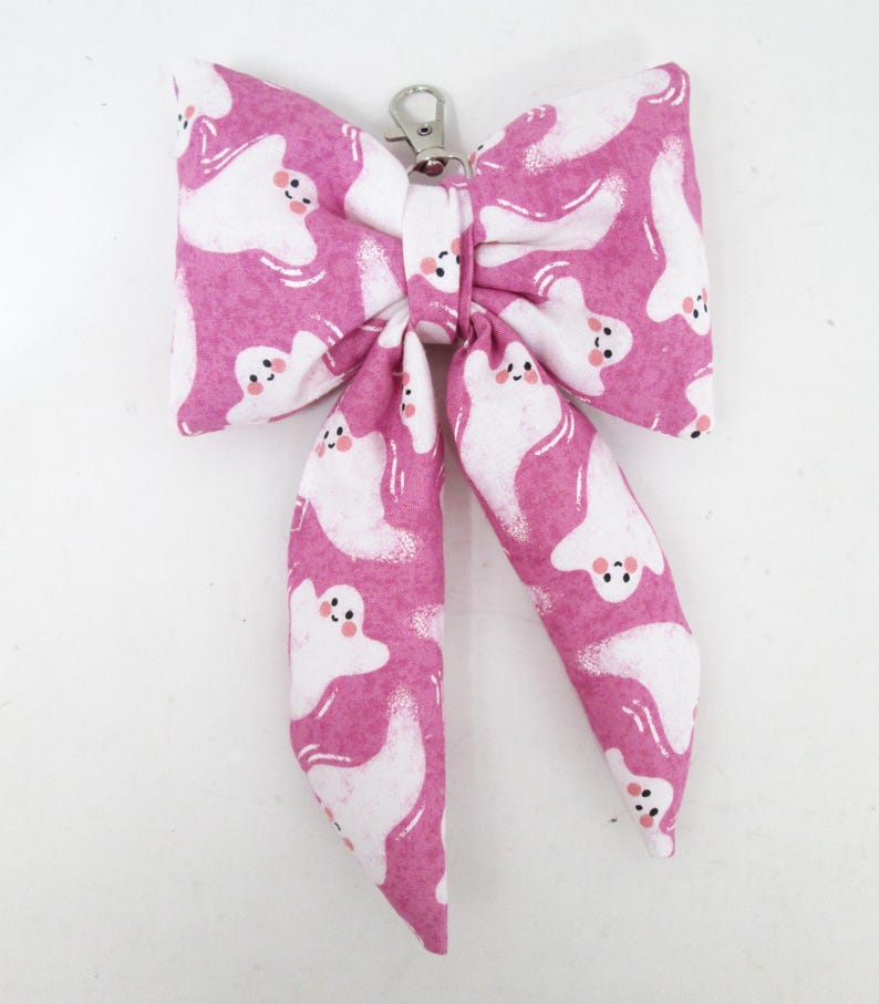 May include: A pink bow-shaped accessory with a white ghost pattern and a silver-colored clasp. The bow is made of fabric and features a repeating design of friendly-looking ghosts. The accessory is likely for a pet or as a decorative item.