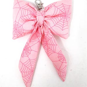 May include: A pink fabric bow with a spiderweb pattern and a silver-colored clasp. The bow is tied in the center and has two long, flowing tails. The spiderweb design is a darker shade of pink.