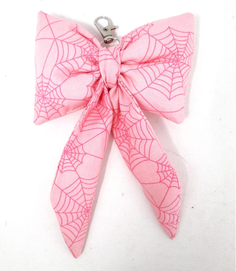 May include: A pink fabric bow with a spiderweb pattern and a silver-colored clasp. The bow is tied in the center and has two long, flowing tails. The spiderweb design is a darker shade of pink.