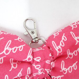 May include: A pink fabric item with the word "boo" in white script repeated across the surface. A silver-colored metal clasp is attached to the fabric. The item appears to be a small accessory or part of a larger product.