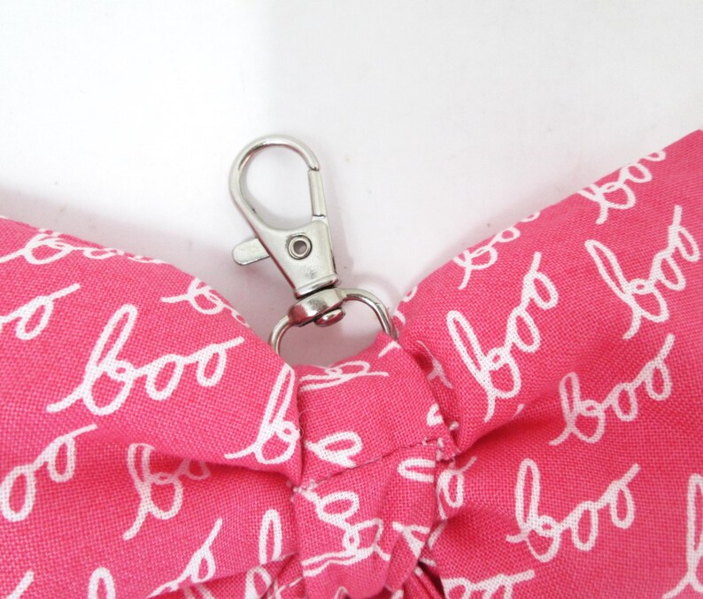 May include: A pink fabric item with the word "boo" in white script repeated across the surface. A silver-colored metal clasp is attached to the fabric. The item appears to be a small accessory or part of a larger product.