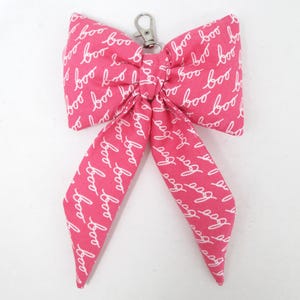 May include: A pink fabric bow with a white "boo" pattern, attached to a silver-colored metal clasp. The bow has long, flowing tails and is suitable for various crafting projects or as a decorative accessory.