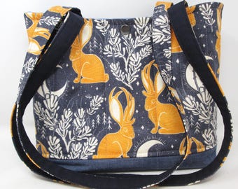 May include: A navy blue tote bag with a repeating pattern of orange rabbits, white foliage, and crescent moons. The bag has a denim base and navy blue straps. The bag is approximately 12 inches wide and 10 inches tall.