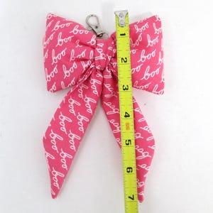 May include: A pink fabric bow with the word "boo" in white script repeated across the surface. The bow is approximately 7 inches long and has a silver-colored metal clasp.