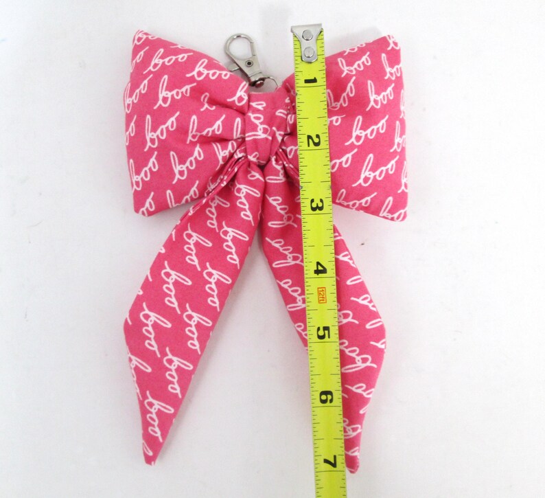 May include: A pink fabric bow with the word "boo" in white script repeated across the surface. The bow is approximately 7 inches long and has a silver-colored metal clasp.