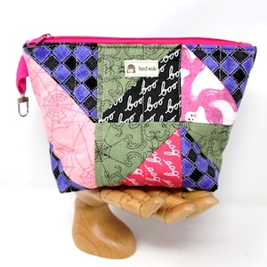 May include: A quilted zippered pouch featuring a patchwork design with purple and black diamond patterns, pink spiderwebs, green scrollwork, black fabric with white 'boo' text, and pink fabric with white ghost shapes. A small 'hand made' label is attached.