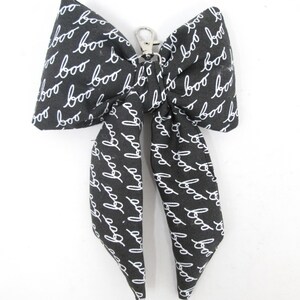 May include: A black and white fabric bow with the word "boo" in a handwritten style repeated across the surface. The bow has a silver-colored metal clasp.