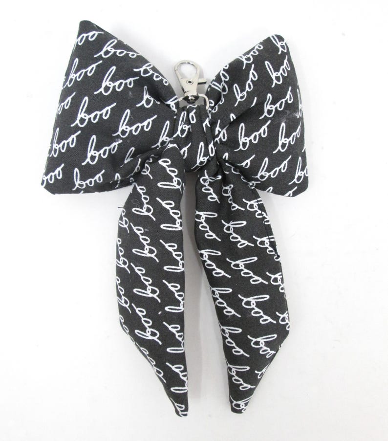 May include: A black and white fabric bow with the word "boo" in a handwritten style repeated across the surface. The bow has a silver-colored metal clasp.