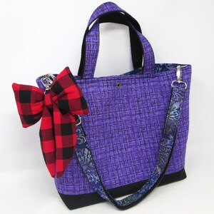 May include: A purple tote bag with black accents and a red and black plaid bow. The bag has a patterned strap and handles. The bag is made of fabric and has a rectangular shape.