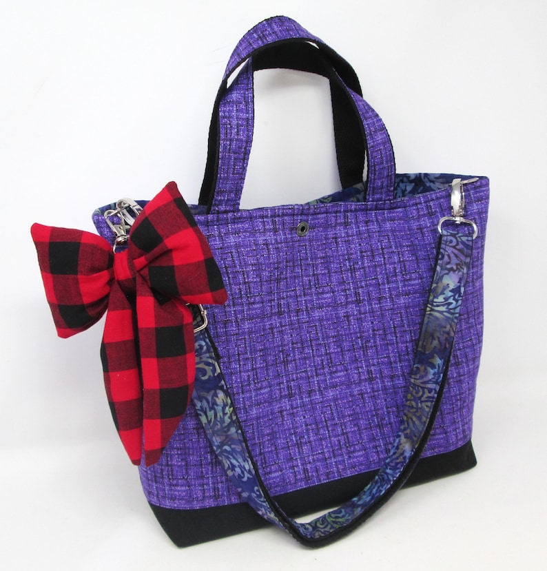May include: A purple tote bag with black accents and a red and black plaid bow. The bag has a patterned strap and handles. The bag is made of fabric and has a rectangular shape.