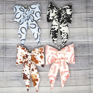 May include: Four fabric bow keyrings with metal clasps. The bows display different patterns: blue with horseshoes, black and white cow print, brown and white cow print, and pink with a western design.
