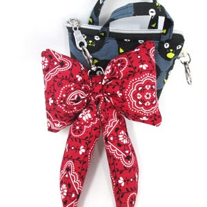 May include: A small, blue tote bag with a black cat pattern and a red bandana bow. The bow features a white paisley design. The bag has a silver zipper and handle. The bow is attached with a silver clasp.
