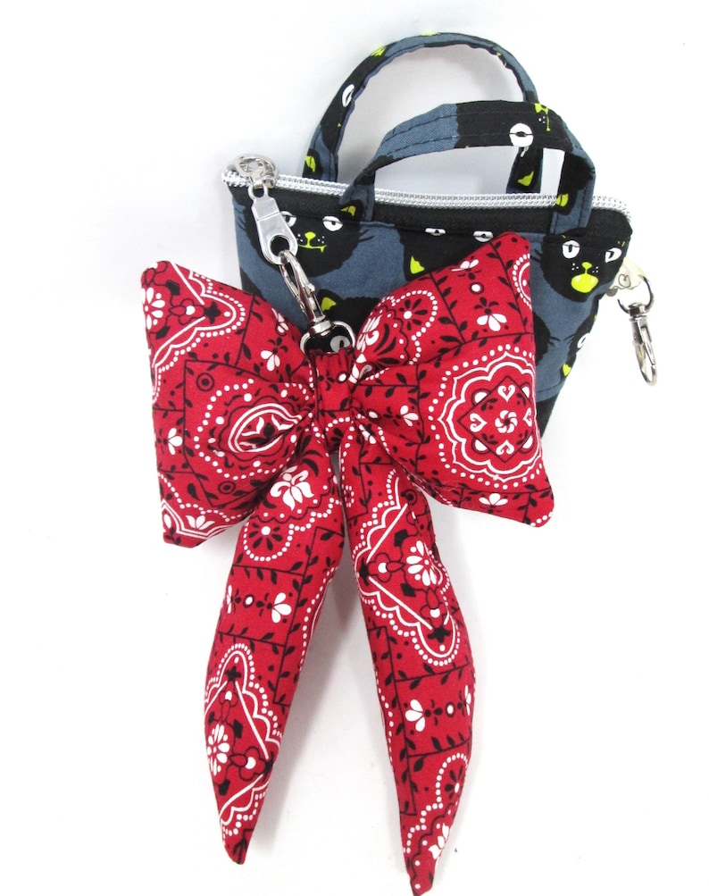 May include: A small, blue tote bag with a black cat pattern and a red bandana bow. The bow features a white paisley design. The bag has a silver zipper and handle. The bow is attached with a silver clasp.