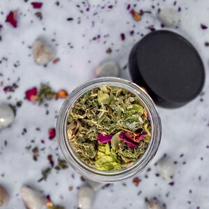 May include: A glass jar filled with a blend of dried herbs and flowers, including green leaves, pink petals, and white buds. The jar is sitting on a white surface with scattered rose petals.