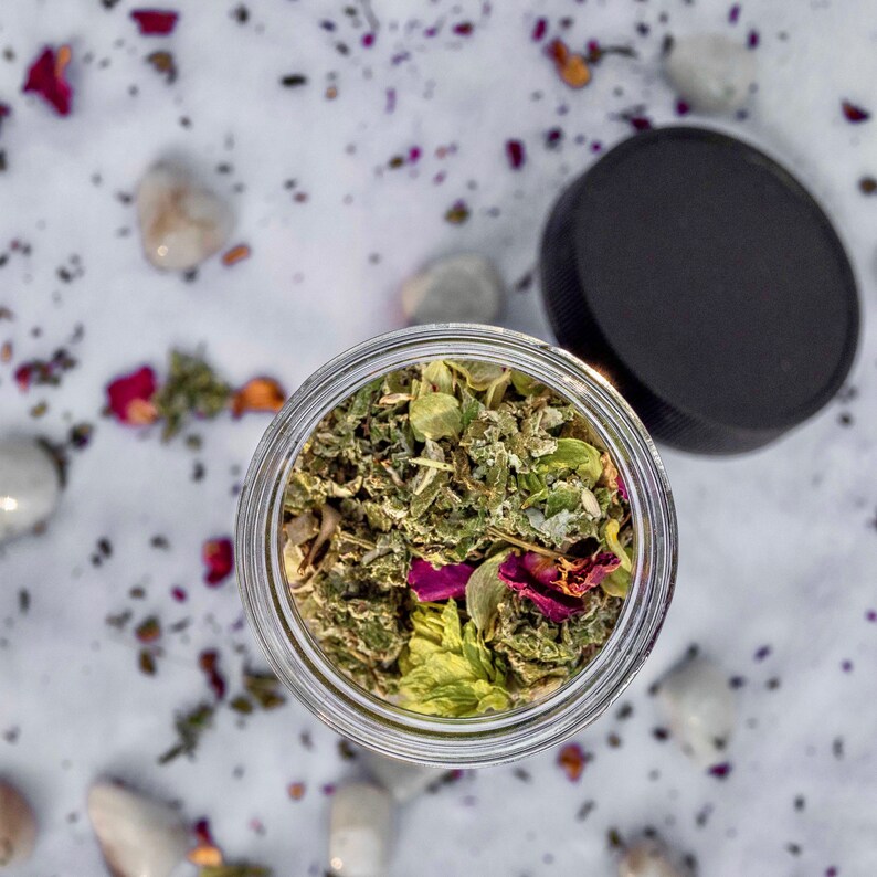 May include: A glass jar filled with a blend of dried herbs and flowers, including green leaves, pink petals, and white buds. The jar is sitting on a white surface with scattered rose petals.